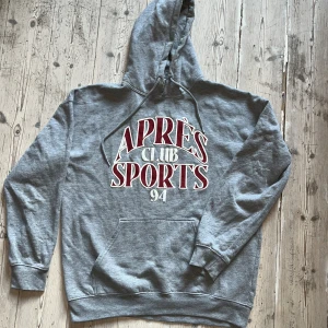 Grå hoodie från Hoxton med tryck - Säljer en grå hoodie från Hoxton med stor vit och vinröd text 'APRÈS CLUB SPORTS 94' på bröstet. Klassisk känguruficka, huva med snörning och mjukt material. Perfekt för chill dagar eller när du vill ha en sportig vibe.