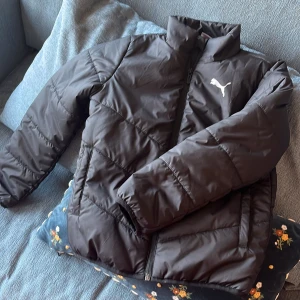 Svart pufferjacka från Puma - Svart pufferjacka för barn storlek S /128 7-8 år från Puma med hög krage och dragkedja framtill. Jackan har ett stilrent snitt och är vadderad för extra värme. Puma-loggan i vitt på bröstet ger en sportig vibe.