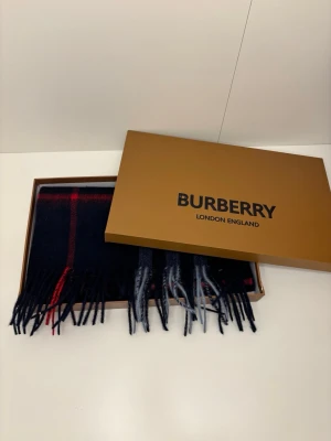 Mörkblå rutig halsduk från Burberry - Snygg mörkblå halsduk från Burberry med klassiskt rutmönster i rött och grått. Halsduken har fransar i ändarna och levereras i originalförpackning med etiketter och certifikat. Perfekt accessoar för att lyfta din stil.