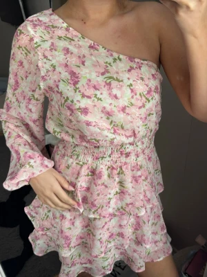 Volang klänning - Söt klänning med blommigt mönster i rosa, vitt och grönt. Klänningen har oneshoulder-design med en lång ärm, volangdetaljer och smock i midjan för en snygg passform. Perfekt till sommaren! 