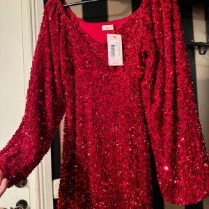 Röd paljettklänning från NA-KD - Glittrig röd klänning från NA-KD med paljetter över hela plagget. Klänningen har lång ärm och v-ringning, perfekt för dig som vill sticka ut. Omlottmodell som ger en snygg siluett.