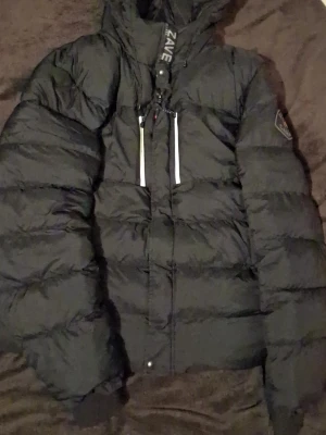 Svart dunjacka från Zavetti - Svart dunjacka från Zavetyi med huva och dragkedja framtill. Jackan har två vertikala fickor med vita detaljer och en patch på ärmen. Perfekt för kalla dagar och har en klassisk pufferstil. XL kan byttas ut mot en burberry jacka helst❤️❤️