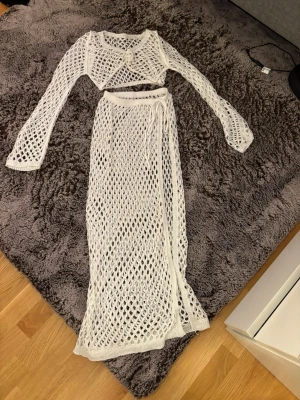Vit stickad strandset i två delar - Snyggt strandset i vitt med långärmad topp och lång kjol i stickat nätmönster. Kjolen har knytband i midjan för perfekt passform. Perfekt att slänga på sig över bikinin på stranden eller vid poolen.