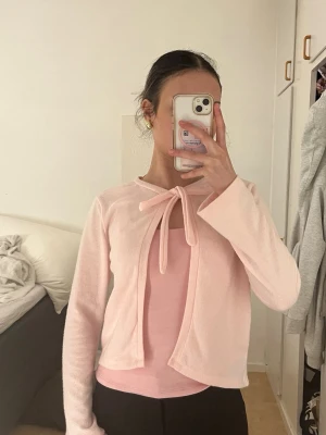 Ljusrosa kofta med rosett - Söt ljusrosa kofta med knytband i halsen och långa ärmar. Koftan har en öppen design framtill och är gjord i ett mjukt, tunt material som passar perfekt till lager-på-lager. Enkel och stilren look.