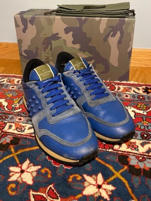 Blå Valentino Rockrunner sneakers - Säljer ett par snygga blå Valentino Rockrunner  med stilrena nitar på sidan. Skorna har blå snörning, rund tå och platt sula. (Det är liiiite slitage på hälen i den högra skon annars är skorna i mycket bra skick). Skorna är perfekta för dig som vill sticka ut med stilrena och exklusiva skor!💙