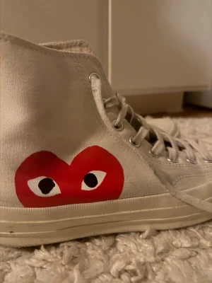 Converse x CDG Play vita high tops - Snygga vita Converse Chuck Taylor high tops i samarbete med Comme des Garçons Play. Skorna har det ikoniska röda hjärta-trycket med ögon på sidan och klassisk snörning. Tillverkade i canvas med gummisula och rund tå.