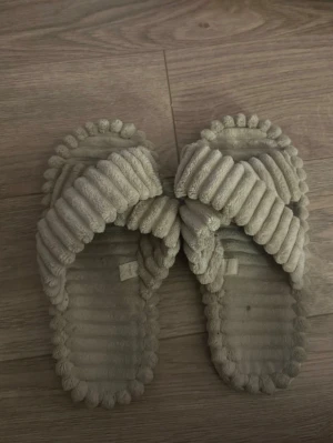 Beige fluffiga tofflor med korsad design - Mysiga beige tofflor med korsad ovandel och ribbad struktur. Tillverkade i mjuk frotté som känns skönt mot fötterna. Perfekta för att glida runt hemma och hålla sig varm om fötterna.