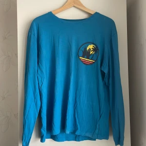 Blå långärmad tröja Summer Camp - Blå långärmad tröja med tryck fram och stort färgglatt motiv på ryggen med texten 'High Five Summer Camp', palmer och solnedgång. Skön passform och perfekt för chill dagar eller häng med vänner. Rund halsringning och mjukt bomullsmaterial.