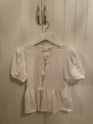 Vit blus med puffärm och knyt - Supersöt vit blus med puffärmar och tre knyt framtill. Blusen har en lätt volang nertill och är tillverkad i mjuk bomull. Perfekt för dig som gillar romantisk och trendig stil.
