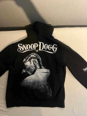 Svart Snoop Dogg hoodie med tryck - Svart hoodie med stor Snoop Dogg-porträtt och logga i vitt på ryggen. Ärmen har tre Snoop Dogg-tryck i vitt. Klassisk huva och ribbade muddar. Perfekt för dig som gillar streetwear och vill sticka ut. Troligtvis i bomullsmix.