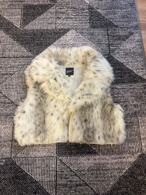 Beige fluffig pälsväst  - Supermjuk och fluffig pälsväst i beige och ljusbruna toner. Västen har en kort, boxig passform och stängs med dragkedja framtill. Perfekt för att addera lite extra stil till din outfit.