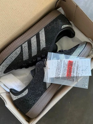 Adidas Samba med strassdetaljer - Snygga Adidas Samba sneakers i svart med glittrande strass över hela skon och klassiska vita ränder på sidorna. Skorna har svart sula och svarta skosnören. Perfekta för dig som vill sticka ut med en unik och trendig look.