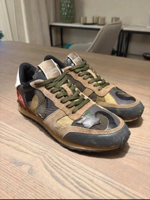 Valentino Rockrunner camo sneakers - Säljer ett par Valentino Rockrunner sneakers med camouflage-mönster i grönt, brunt och svart. Skorna har detaljer i mocka, skinn och röd textil på hälen samt gröna snören och en platt sula. Perfekta för dig som vill sticka ut med en unik design.