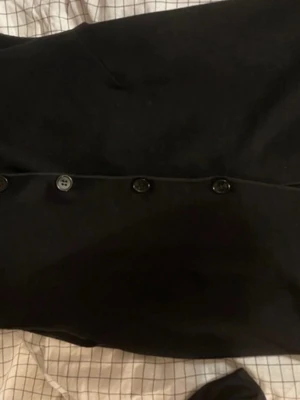 Svart väst med knappar - Stilren svart väst med knäppning framtill. Västen är ärmlös och har en rak passform som passar perfekt till lager-på-lager-stil. Materialet ser ut att vara mjukt tyg, vilket gör den bekväm att bära. Enkel design utan synliga loggor eller mönster.