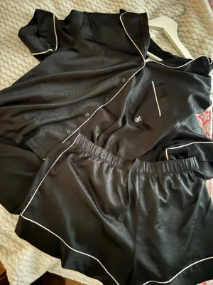 Svart satinpyjamas med vita detaljer - Snygg svart pyjamas från Victoria's Secret i glansigt satinmaterial. Setet består av en kortärmad skjorta med vita passpoaler och matchande shorts med elastisk midja. Diskret logotyp på bröstfickan och ton-i-ton-mönster i tyget.