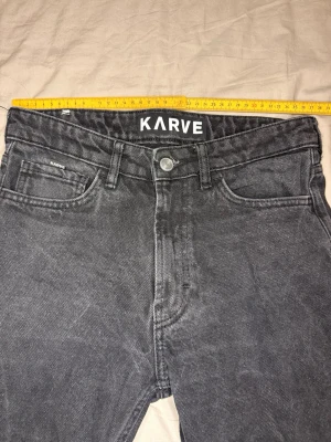 Mörkgrå jeans från KARVE - Säljer ett par mörkgrå jeans från KARVE med klassisk femficksdesign och knappgylf. Jeansen har en rak passform och är tillverkade i ett slitstarkt bomullsmaterial. Perfekta för dig som gillar en enkel och stilren look.