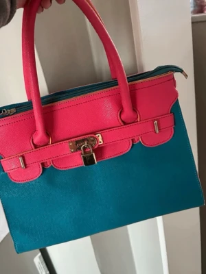 Blå och rosa handväska med låsdetalj - Färgstark handväska i blått och rosa med guldfärgade metalldetaljer och dekorativt lås framtill. Väskan har dubbla handtag och dragkedja upptill. Perfekt för dig som vill sticka ut med din stil.