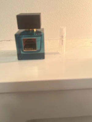 Blå fyrkantig parfymflaska rituals - Snygg parfymflaska i blått glas med svart fyrkantigt lock och gulddetaljer. Flaskan har en modern, stilren design och kommer med ett litet provrör. Perfekt för dig som gillar exklusiva dofter och vill ha en elegant flaska på hyllan.