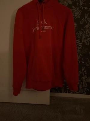 Röd hoodie från Peak Performance - Snygg röd hoodie från Peak Performance med broderad logga framtill och klassisk känguruficka. Hoodien har dragsko i huvan och ribbade muddar vid ärmslut och nederkant. Perfekt för en avslappnad och sportig stil.