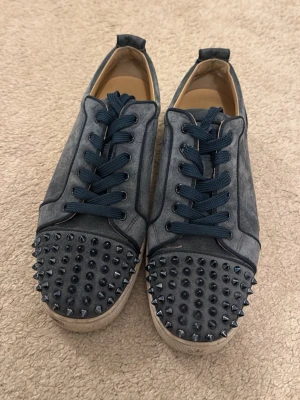Louboutin - Unika blåa sneakers från Christian Louboutin med blåa nitar på tån, blåa snören och den klassiska röda sulan. Skorna har en rund tå och är tillverkade i mocka för en lyxig känsla. Perfekta för dig som vill sticka ut med din stil. Kan även tvättas vid köp