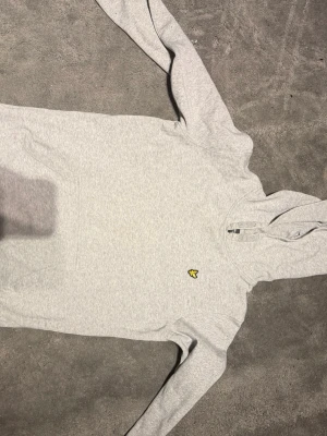 Grå hoodie från Lyle & Scott Junior - Ljusgrå hoodie från Lyle & Scott Junior med klassisk huva och lång ärm. Tröjan har det ikoniska gula örnlogot på bröstet och är gjord i mjukt bomullsmaterial. Enkel och stilren design som funkar till det mesta.