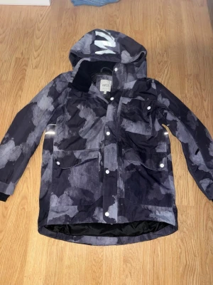 Mönstrad regnjacka från Didriksons - Säljer en snygg regnjacka från WACC med grått och svart abstrakt mönster. Jackan har huva, flera fickor med knappar och reflexdetalj på ryggen. Perfekt för regniga dagar och har en modern look.