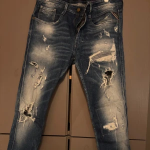 Replay Anbass blå slitna jeans 33/32 - Säljer ett par Replay Anbass jeans i blå tvätt med coola slitningar och trasiga detaljer framtill. Jeansen har klassisk femficksdesign, normal passform och är gjorda i jeansmaterial. Perfekt för dig som gillar en edgy streetstil.