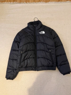 Svart pufferjacka från The North Face - Snygg svart pufferjacka från The North Face med hög krage och dragkedja framtill. Jackan har två fickor med dragkedja och logga på bröstet och ryggen. Perfekt för kalla dagar och ger en cool, avslappnad look.Jacka, ny, använd bara 2–3 gånger.