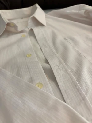 Vit randig skjorta med knappar - Klassisk vit skjorta med diskreta vertikala ränder och gulaktiga knappar. Skjortan har lång ärm, bröstficka och en stilren krage. Perfekt för dig som gillar en clean och enkel look. Materialet känns som bomull och passformen är normal.