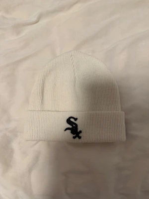 Vit ribbad mössa New Era Sox - Säljer en stilren vit ribbad mössa från New Era med Chicago White Sox-logga broderad i svart framtill. Mössan har uppvikt kant och är gjord i mjukt, stretchigt material som sitter skönt på huvudet. Perfekt för dig som gillar sportig streetstyle.