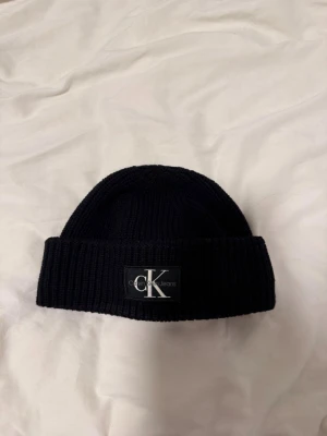 Svart ribbstickad mössa Calvin Klein - Svart ribbstickad mössa från Calvin Klein Jeans med uppvikt kant och tydlig CK-logga framtill. Klassisk och stilren design som passar perfekt till streetwear eller en enkel vardagslook. Materialet är mjukt och håller värmen under kalla dagar.
