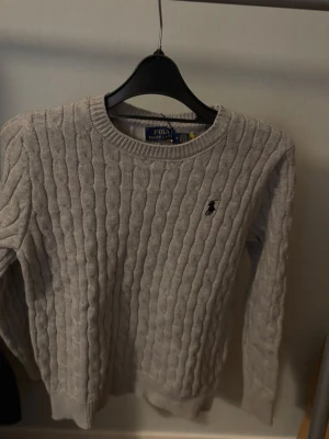 Grå kabelstickad tröja Polo Ralph Lauren - Snygg grå kabelstickad tröja från Polo Ralph Lauren med rund halsringning och klassisk broderad logga på bröstet. Tröjan har långa ärmar och ribbade muddar vid ärmslut och nederkant. Perfekt för lager på lager och stilrena outfits.