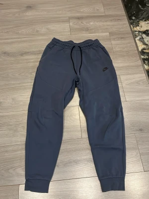 Mörkblå mjukisbyxor från Nike - Snygga mörkblå mjukisbyxor från Nike med svart snörning i midjan och diskret Nike-logga på vänster ben. Byxorna har muddar vid bensluten och är perfekta för chill eller träning. Tillverkade i mjukt material för maximal komfort.