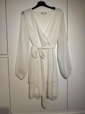 Vit omlottklänning med knytband - Söt vit kortklänning med omlottmodell och knytband i midjan. Klänningen har långärmade, lätt transparenta ärmar och diskret prickigt mönster i tyget. Volangdetaljer nedtill ger en extra touch. Perfekt för sommarens alla tillfällen!