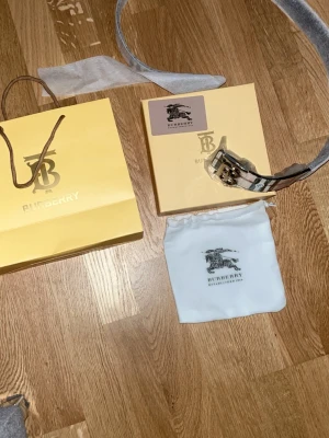 Burberry bälte med silver logo - Snyggt Burberry-bälte i klassiskt beige rutmönster med ett stort, silverfärgat TB-spänne. Tillverkat i läder och levereras med originalpåse och ask. Perfekt accessoar för att lyfta vilken outfit som helst. (2 sidig, svart och original)