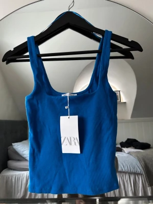 Blå ribbad linnetopp från Zara - Snygg blå ribbad linnetopp från Zara med breda axelband och rundad halsringning. Storlek S men passar XS. Toppen är croppad och har en tight passform, perfekt att styla till jeans eller kjol. Tillverkad i mjuk bomullsmix för skön känsla.