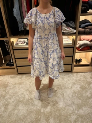 Blommig vit och blå kortklänning - Söt kortklänning med volangärmar och blommigt mönster i vitt och ljusblått. Klänningen har en luftig passform och rund halsringning, perfekt för varma dagar. Tillverkad i ett lätt och bekvämt material som ger en flowy känsla. Rekommenderar att testa snurra i den! Hahaha