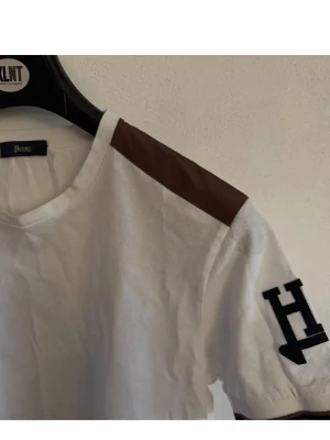 Herno T-shirt  - Snygg vit t-shirt från Herno med bruna axelpartier och stort H-tryck på ärmen. Klassisk rund halsringning och korta ärmar. Perfekt för dig som gillar stilrena detaljer och vill ha något unikt i garderoben.