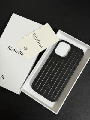 Rimowa iPhone 17 svart mobilskal - Stilrent svart mobilskal från Rimowa med räfflad design och diskret logotyp. Skalet är tillverkat i tåligt material och har en robust känsla. Passar iPhone och ger ett exklusivt intryck med sin eleganta finish och skyddande kanter runt kameran.