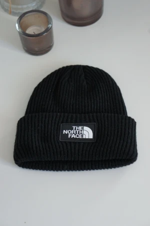 Svart ribbad mössa The North Face - Stilren svart ribbstickad mössa från The North Face med uppvikt kant och tydlig logotyp framtill. Klassisk passform och mjukt material som håller dig varm under kalla dagar. Perfekt accessoar för en enkel och trendig look.
