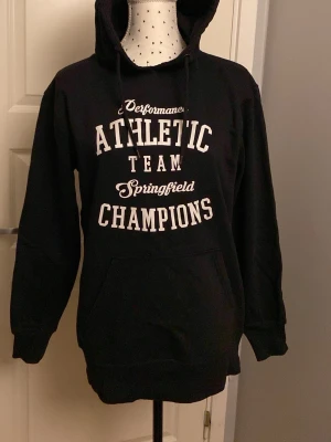 Svart hoodie Springfield Athletic - Svart hoodie med vit text 'Performance Athletic Team Springfield Champions' på bröstet. Klassisk känguruficka framtill, huva med dragsko och ribbade muddar. Perfekt för en sportig och avslappnad stil.