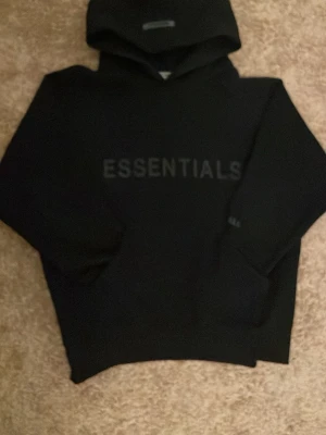 Svart Essentials Fear of God hoodie - Snygg svart hoodie från Fear of God Essentials med stor logga på bröstet och mindre tryck på ärmen. Klassisk passform med huva och ribbade muddar. Tillverkad i mjukt bomullsmaterial som känns riktigt skönt mot huden. Kom gärna med bud!!