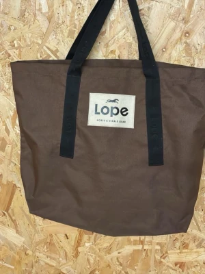Brun shoppingväska från Lope - Stor brun shoppingväska från Lope med svarta handtag och en tydlig logga framtill. Väskan är rymlig och har en enkel, stilren design som passar perfekt för att bära med sig allt man behöver under dagen.