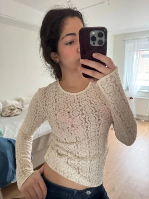 Vit spetstopp med lång ärm - Säljer en vit, långärmad blus i genombruten spets med blommigt mönster. Toppen har en rund halsringning och är figurnära i modellen. Perfekt för dig som gillar romantiska och trendiga plagg.