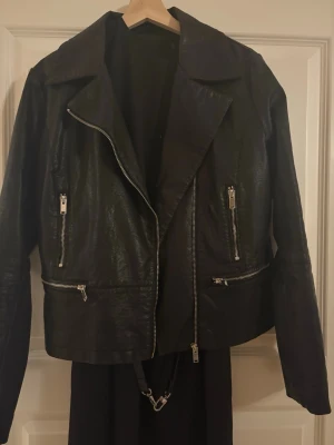 Svart skinnjacka med dragkedjor - Snygg svart skinnjacka med flera silverfärgade dragkedjor och klassisk krage. Jackan har lång ärm och en rak, något kortare passform. Perfekt för dig som vill ha en stilren och edgy look. Str 38/M. pris 150kr