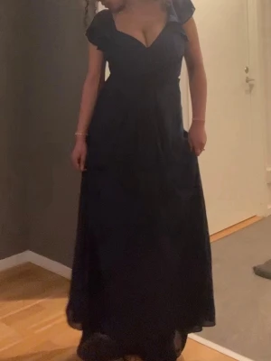 Marinblå långklänning i polyester - Snygg marinblå långklänning med ärmlös design och klassisk siluett. Klänningen har en elegant och enkel look, perfekt för speciella tillfällen som balen. - Material utan stretch - Foder - Avskärning i midjan - Avtagbart knytband - Dold ryggdragkedja - Drapering över bysten och vid midjan  Material Yttertyg: 100% polyester av återvunnen fiber. Foder: 100% polyester av återvunnen fiber. Tvättråd  Tvätt 30°.