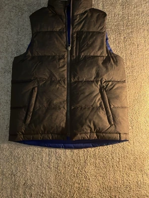 Svart dunväst från Polo Ralph Lauren - Stilren svart dunväst från Polo Ralph Lauren med diskret broderad logga på bröstet. Västen har dragkedja framtill, två snedställda fickor och blått innerfoder. Perfekt för lager-på-lager och håller dig varm under kyliga dagar. Dubbel sidig 9/10 skick