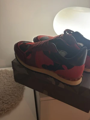 Valentino Garavani röda sneakers - Snygga sneakers från Valentino Garavani i rött och svart med camouflage-mönster. Skorna har snörning, rund tå och platt sula. Materialmix av skinn och textil ger en lyxig känsla. Perfekta för dig som vill sticka ut med exklusiva detaljer.
