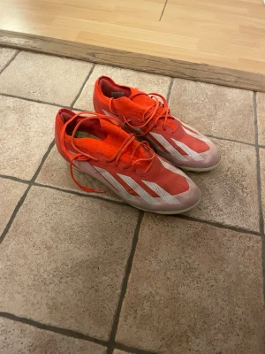Röda Adidas fotbollsskor med snörning - Säljer ett par röda Adidas fotbollsskor med vita detaljer och snörning. Skorna har en låg profil och är tillverkade i syntetmaterial för bra grepp och känsla på planen. Perfekta för dig som vill sticka ut på fotbollsplanen.