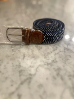 Blått flätat bälte med läderdetaljer - Snyggt blått flätat bälte med silverfärgat spänne och bruna läderdetaljer vid ändarna. Bältet har ett klassiskt mönster och passar perfekt till jeans eller chinos. Materialet är en mix av textil och läder för både stil och komfort.